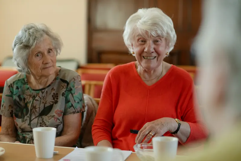 Twee oudere vrouwen zitten lachend aan tafel met een kop koffie tijdens een koffieochtend in een buurthuis.